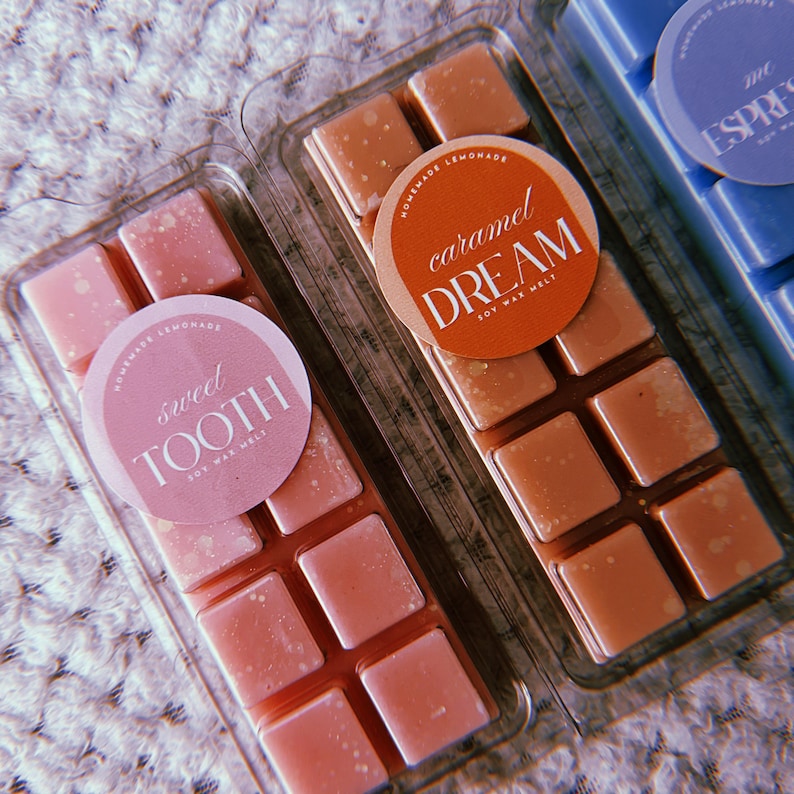 May include: Two packs of soy wax melts in clear plastic packaging. The wax melts are in a grid of six squares. The left pack is pink and has a white circle label that says "sweet tooth soy wax melt". The right pack is brown and has a white circle label that says "caramel dream soy wax melt".