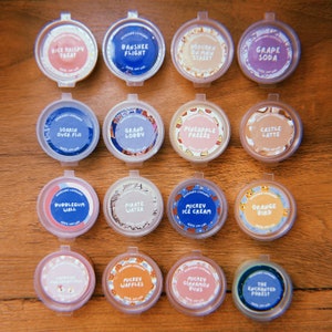 May include: Sixteen circular containers with lids, each labelled with a different name and design. The labels include names like "Rice Krispy Treat", "Banshee Flight", and "Grape Soda". The containers are arranged on a wooden surface.