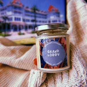 May include: A glass jar candle with a white label that says "Homemade Lemonade Grand Lobby Magical Soy Candles". The candle is in a cozy setting with a knitted blanket.