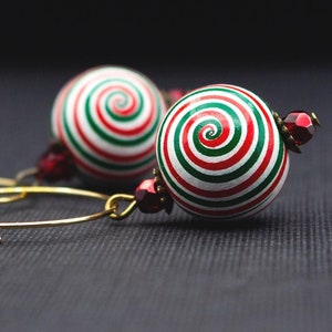 Könnte beinhalten: Goldene Creolen mit kugelförmigen Perlen mit einem Spiralmuster in Rot, Grün und Weiß. Kleine, dunkelrote Perlen sind ebenfalls vorhanden. Die Ohrringe sind vor einem dunklen Hintergrund platziert.