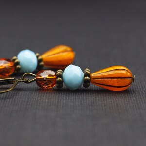 Könnte beinhalten: Nahaufnahme von Ohrringen im Vintage-Stil. Die Ohrringe bestehen aus orangefarbenen, hellblauen und bernsteinfarbenen Perlen. Das Design umfasst eine tropfenförmige orangefarbene Perle und einen Klappbügelverschluss.
