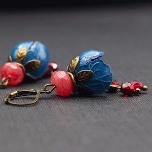Könnte beinhalten: Ein Paar blaue und rote Ohrringe mit einem floralen Design. Die Ohrringe haben eine blaue Blume mit goldenen Akzenten und einen roten Edelstein.