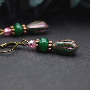 May include: A pair of dangle earrings featuring green and pink beads with bronze-colored accents. The earrings have a teardrop-shaped bead with a metallic finish and a lever-back closure. The design includes small pink beads and a larger green bead.