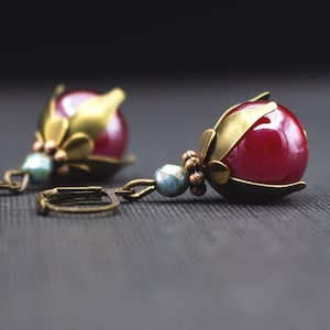 以下が含まれることがあります： 深紅色の球形ビーズを金色の金属細工で囲んだ、装飾的なイヤリングのペア。イヤリングは、小さな水色のビーズとアンティーク調の留め金が付いた花のモチーフで、暗い背景にセットされています。