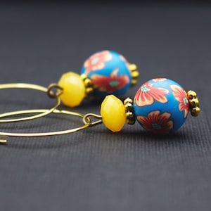 Könnte beinhalten: Ein Paar goldene Creolen mit blauen Blumenperlen und gelben facettierten Perlen. Die blauen Perlen weisen ein rotes und orangefarbenes Blumenmuster auf. Die Ohrringe sind vor einem dunkelgrauen Hintergrund platziert.