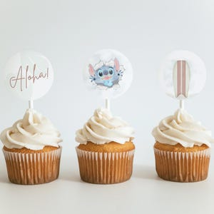 May include: Three cupcakes with white frosting topped with round paper circles. The circles have images of a cartoon character, a surfboard, and the word "Aloha!" in pink script.