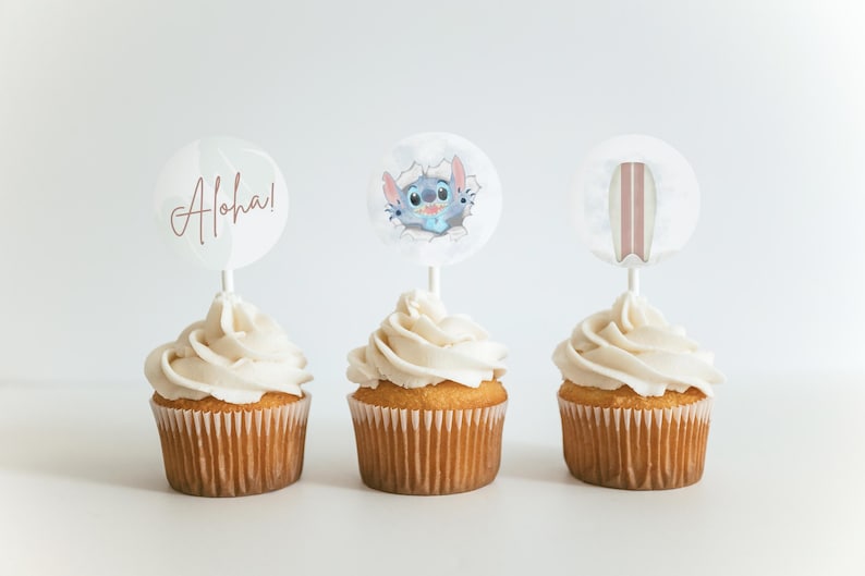 May include: Three cupcakes with white frosting topped with round paper circles. The circles have images of a cartoon character, a surfboard, and the word "Aloha!" in pink script.