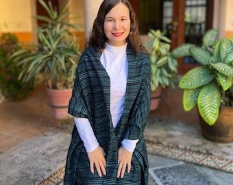 May include: A woman wearing a teal and black striped shawl with black fringe. The shawl is draped over a white long-sleeved shirt. The woman is standing in a courtyard with plants and a stone pillar in the background.