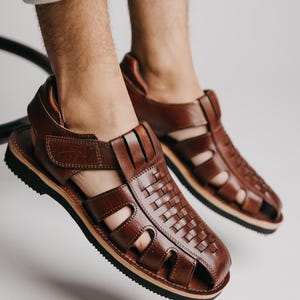 May include: Brown leather sandals with a woven design on the top. The sandals have a black sole and a strap that fastens with a buckle. The sandals are open-toed and appear to be new.