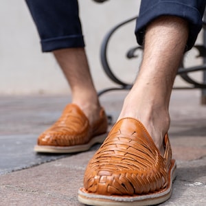 May include: Close-up of brown leather woven sandals with a white sole. The sandals are worn by a person wearing navy blue pants. The sandals have a classic design and are suitable for casual wear.