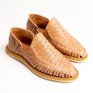 May include: A pair of tan leather woven slip-on shoes. The shoes have a rounded toe and a woven design across the top. The soles are white with a thin orange stripe.