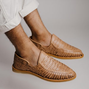May include: Tan leather woven slip-on shoes. The shoes have a rounded toe and a woven design across the top. The soles are a light brown color. The shoes are shown on a person wearing white pants.