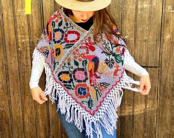 May include: A gray poncho with white fringe and colorful embroidered flowers, birds, and geometric patterns. The poncho is worn by a person wearing a brown hat and blue jeans.