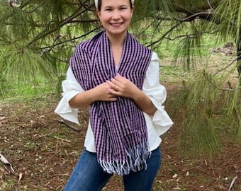 May include: A woman wearing a white shirt and blue jeans is standing in front of a pine tree. She is wearing a purple and white striped scarf with fringe.