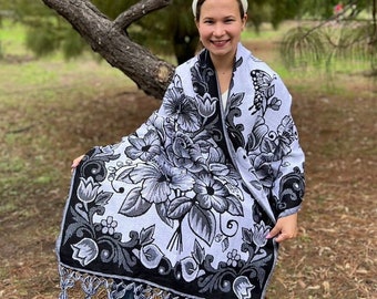 May include: A woman wearing a white headband sits on a tree branch, wrapped in a light blue and black shawl with a floral pattern and black tassels.