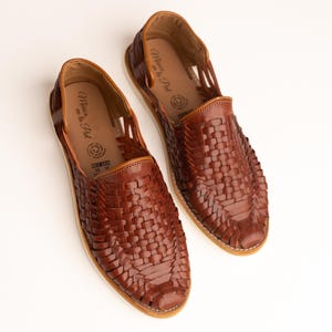 May include: A pair of brown leather woven huarache sandals. The sandals have a slip-on design with a woven upper and a light-colored sole. The interior of the shoe has a printed logo. The shoes are displayed on a white background.