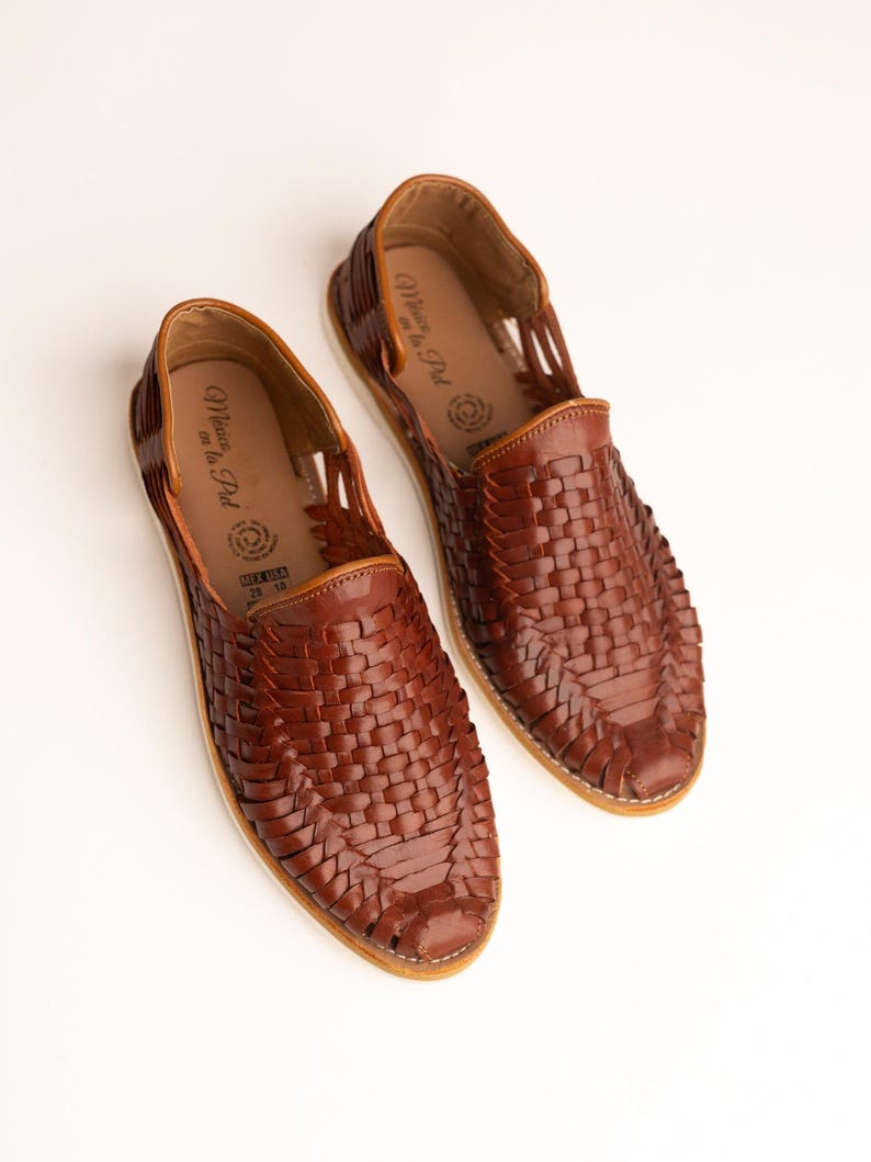 May include: A pair of brown leather woven huarache sandals. The sandals have a slip-on design with a woven upper and a light-colored sole. The interior of the shoe has a printed logo. The shoes are displayed on a white background.