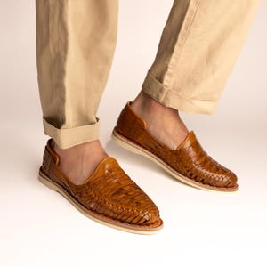 May include: Woven leather shoes in a warm brown hue. The shoes feature a unique open design with a textured, basket-weave pattern. The soles are a light color, and the shoes are paired with tan-colored pants.