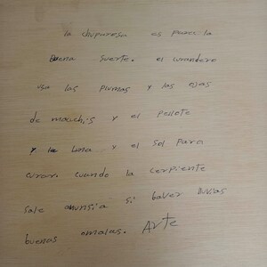 May include: A light-colored wooden surface featuring handwritten black ink text in Spanish that reads: la chuparesa es para la buena suerte. el curandero usa las plumas y las ojas de mauchis y el pellote y la luna y el sol para curar. cuando la serpiente sale anunsi'a si baver lluvias buenas omalas. Arte.