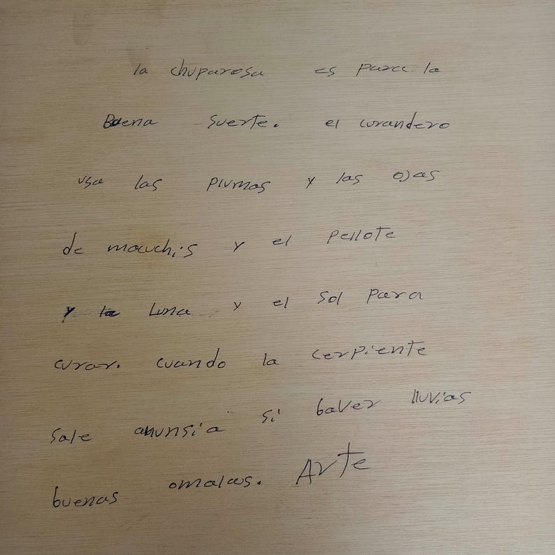 May include: A light-colored wooden surface featuring handwritten black ink text in Spanish that reads: la chuparesa es para la buena suerte. el curandero usa las plumas y las ojas de mauchis y el pellote y la luna y el sol para curar. cuando la serpiente sale anunsi'a si baver lluvias buenas omalas. Arte.