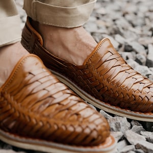 May include: Close-up of brown leather woven shoes. The shoes have a slip-on design with a woven pattern across the top. The soles are white with a brown trim. The shoes are worn with khaki-colored pants.