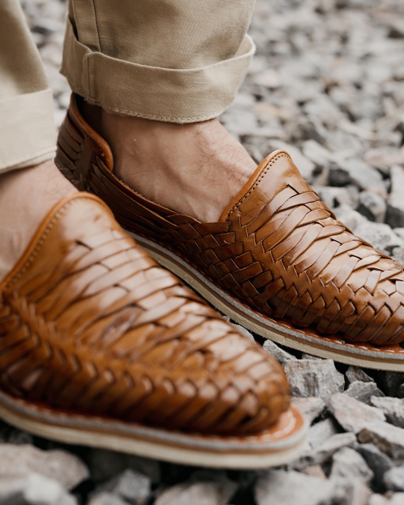 May include: Close-up of brown leather woven shoes. The shoes have a slip-on design with a woven pattern across the top. The soles are white with a brown trim. The shoes are worn with khaki-colored pants.
