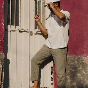 May include: A person wearing a white cap, white short-sleeve shirt, and olive green pants stands near a white door and a magenta wall. The person is also wearing brown woven leather shoes.