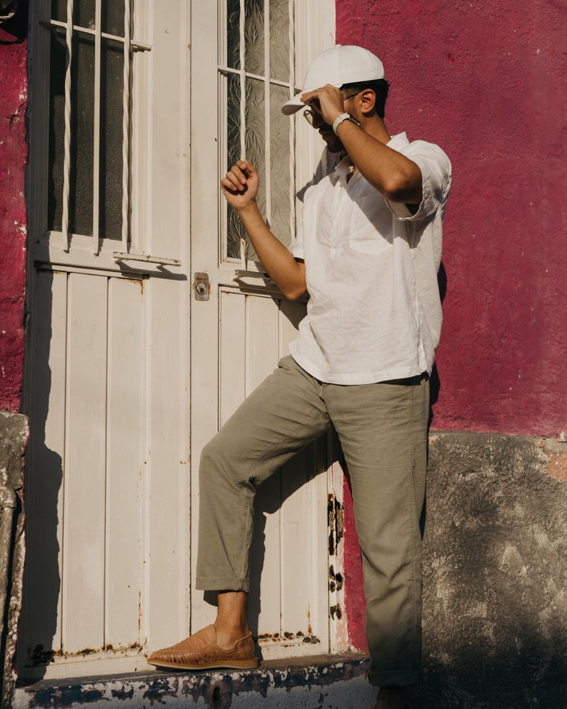 May include: A person wearing a white cap, white short-sleeve shirt, and olive green pants stands near a white door and a magenta wall. The person is also wearing brown woven leather shoes.