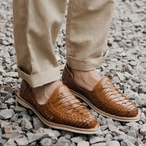 May include: Brown leather woven loafers. The shoes have a slip-on design with a woven upper and a white sole. The loafers are worn with khaki-colored pants. The shoes are made of leather and are designed for casual wear.