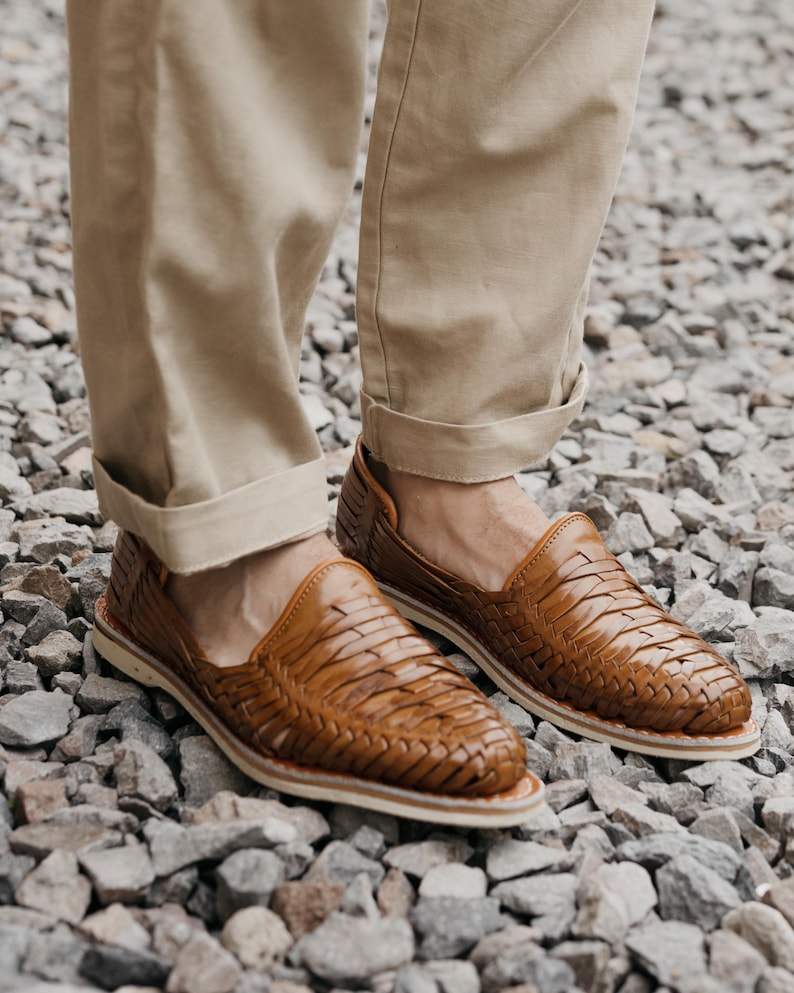 May include: Brown leather woven loafers. The shoes have a slip-on design with a woven upper and a white sole. The loafers are worn with khaki-colored pants. The shoes are made of leather and are designed for casual wear.