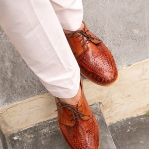 May include: Brown leather shoes with a woven pattern and lace-up closure. The shoes are worn by a person wearing white pants.