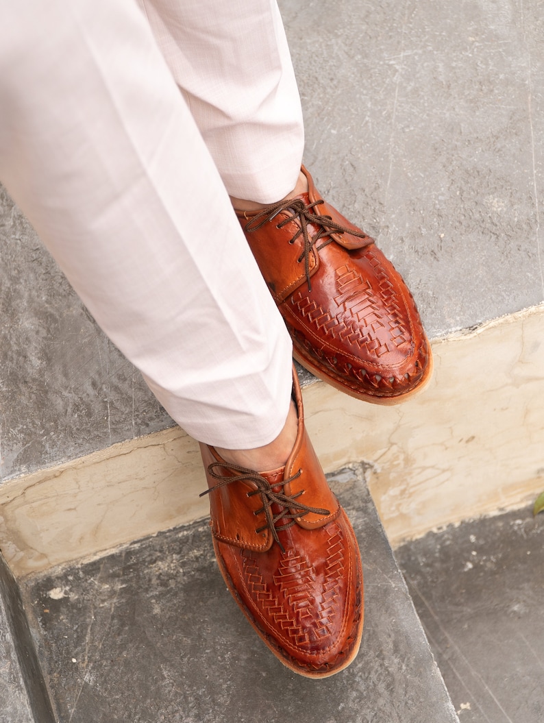 May include: Brown leather shoes with a woven pattern and lace-up closure. The shoes are worn by a person wearing white pants.
