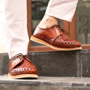 May include: A pair of brown leather shoes with woven detailing. The shoes have laces and are worn by a person standing on a set of stairs.