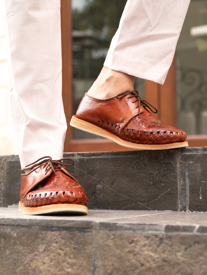 May include: A pair of brown leather shoes with woven detailing. The shoes have laces and are worn by a person standing on a set of stairs.