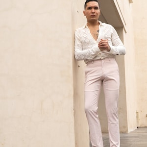 May include: A man wearing a white crocheted shirt with a geometric pattern, light pink pants, and brown leather shoes. He is standing in a doorway with a white wall behind him.