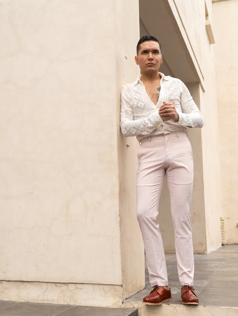 May include: A man wearing a white crocheted shirt with a geometric pattern, light pink pants, and brown leather shoes. He is standing in a doorway with a white wall behind him.
