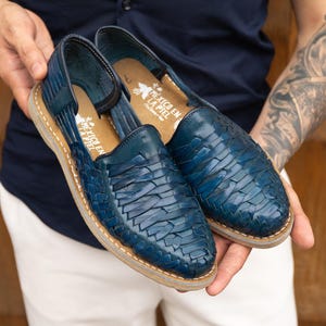 May include: A pair of blue woven leather huarache sandals. The sandals have a woven design on the upper and a tan sole. The text "MEXICO EN LA PIEL" is visible on the insole. The sandals are held in front of a blurred background.