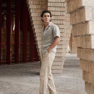 May include: A person wearing a short-sleeved, light green button-down shirt and khaki pants. The person is wearing brown woven leather loafers. The background features brick columns and a red door.
