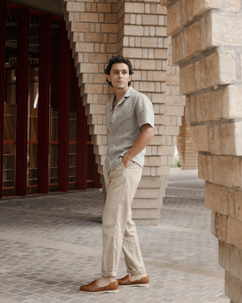 May include: A person wearing a short-sleeved, light green button-down shirt and khaki pants. The person is wearing brown woven leather loafers. The background features brick columns and a red door.