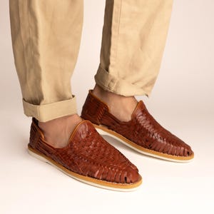 May include: Brown leather woven slip-on shoes with a white sole. The shoes are worn with khaki-colored pants. The shoes have a woven design and a leather upper. The shoes are a casual style.