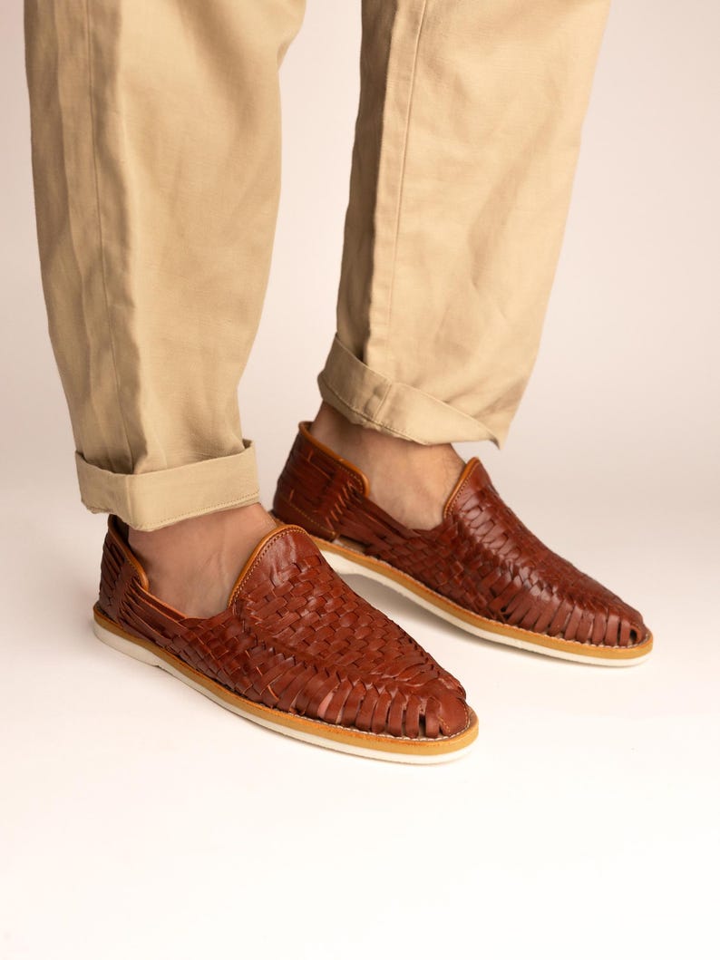 May include: Brown leather woven slip-on shoes with a white sole. The shoes are worn with khaki-colored pants. The shoes have a woven design and a leather upper. The shoes are a casual style.