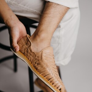May include: Tan leather huaraches with woven detailing. The open-toe sandals have a light-colored sole with a thin yellow stripe. The shoes are being held by a person wearing white pants.