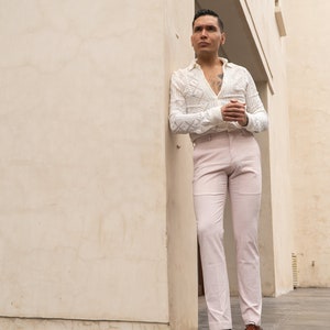 May include: A man wearing a white crocheted shirt with a pink pair of pants and brown leather shoes. He is standing in a doorway with a white wall behind him.