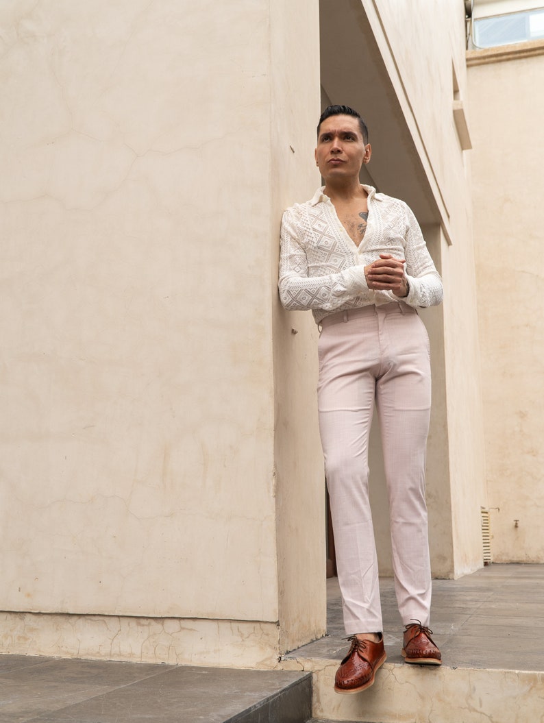 May include: A man wearing a white crocheted shirt with a pink pair of pants and brown leather shoes. He is standing in a doorway with a white wall behind him.