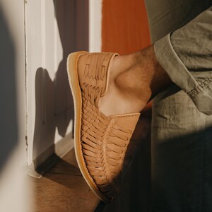 May include: Tan leather woven slip-on shoe with a light-colored sole. The shoe features a unique woven design across the top and sides. The shoe is shown on a person's foot, with the leg wearing olive-green pants.