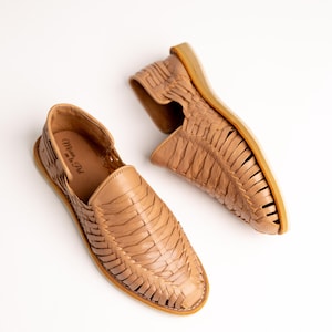 May include: A pair of tan leather huaraches, a traditional Mexican sandal. The sandals feature a woven design across the top and a flat sole. The words "Mexico en la Piel" are visible on the side of one sandal.
