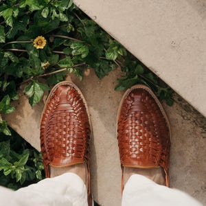 May include: A top-down view of brown leather woven shoes. The shoes are a medium brown color and have a woven pattern across the top. The shoes are on a concrete surface with green plants and a yellow flower in the background.