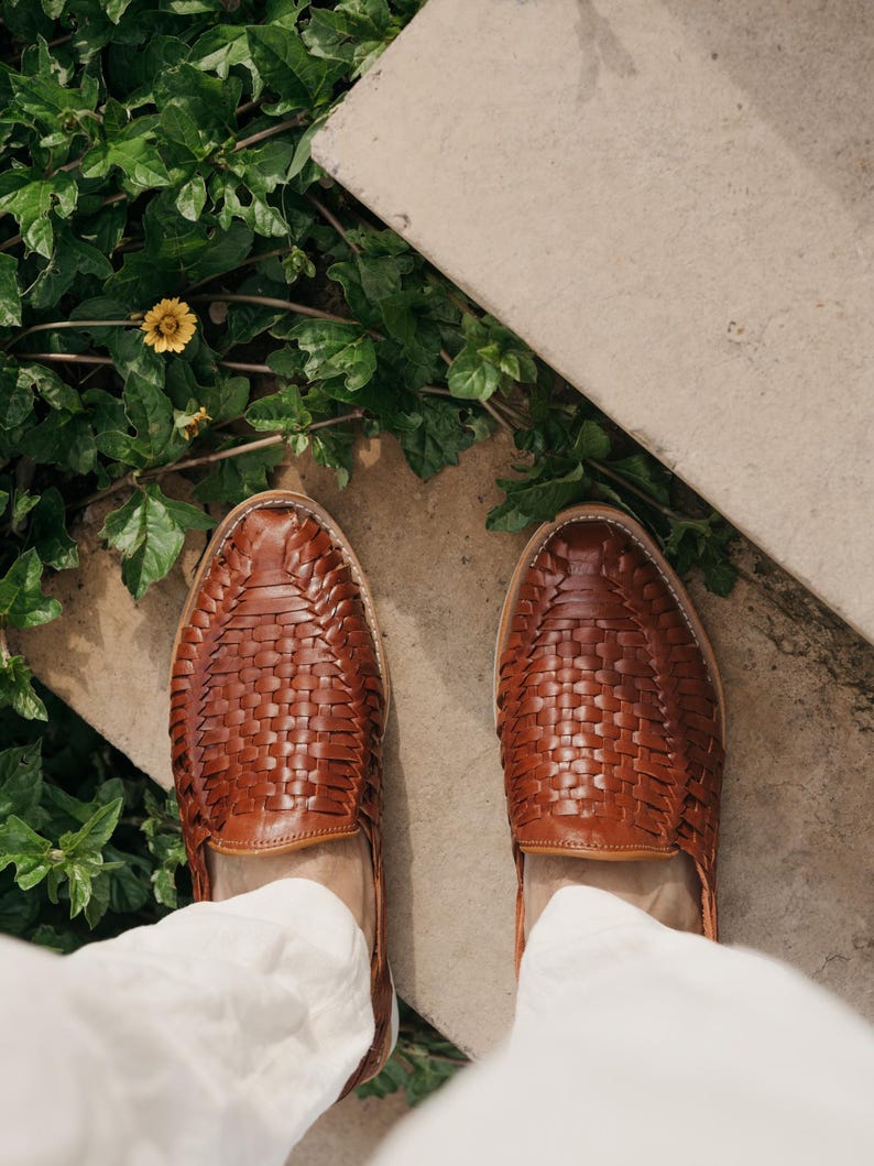 May include: A top-down view of brown leather woven shoes. The shoes are a medium brown color and have a woven pattern across the top. The shoes are on a concrete surface with green plants and a yellow flower in the background.