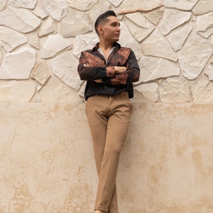 May include: A person wearing a brown and black patterned shirt with a black button-down collar, brown pants, and black shoes. The person is standing in front of a white stone wall.