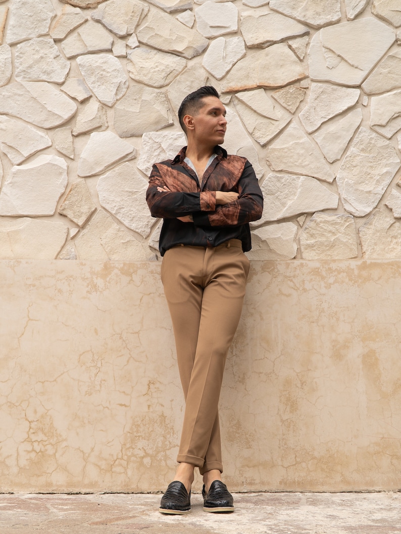 May include: A person wearing a brown and black patterned shirt with a black button-down collar, brown pants, and black shoes. The person is standing in front of a white stone wall.